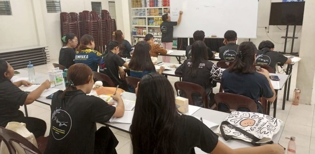 Des étudiants écoutent un enseignant qui écrit sur un tableau blanc dans une salle de classe.