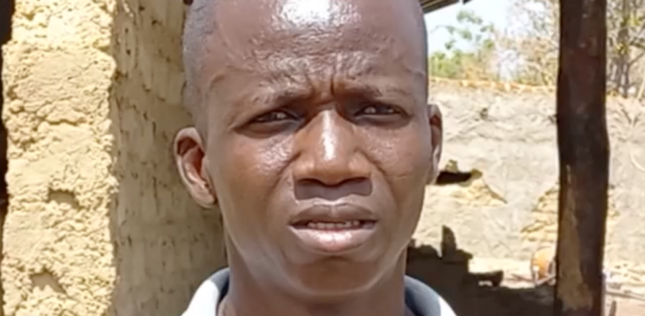 Un homme se tient dehors, face à la caméra. Il semble triste ou préoccupé.