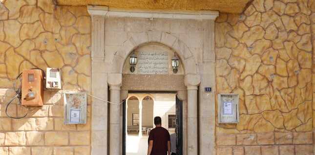 Deux hommes entrent dans un bâtiment en pierre ocre à travers une porte voûtée portant une inscription en arabe.