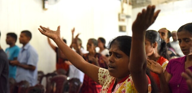ChatGPT a dit :  Une femme au premier plan prie les yeux fermés et les bras levés, entourée d’autres personnes qui prient dans une église.