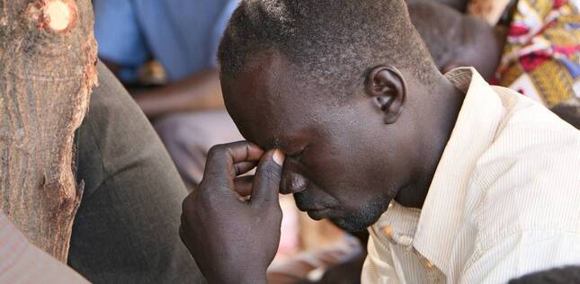 Un homme assis, la tête baissée et les yeux fermés, prie en silence, le visage appuyé contre sa main.