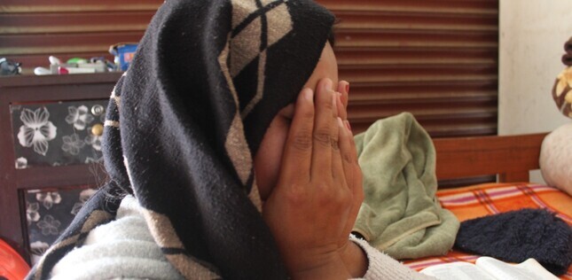Jeune femme la tête couverte d’un châle, le visage caché entre ses mains, avec une Bible ouverte devant elle sur la table.