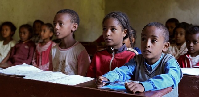 Des enfants sont assis à des pupitres dans une salle de classe, attentifs, avec des cahiers ouverts devant eux.