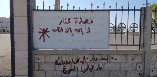 Mur et panneau d’une église tagués de graffitis en arabe, écrits en rouge, devant une clôture métallique.