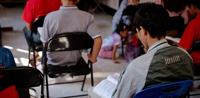 Personnes assises sur des chaises, lisant des livres ou des documents, dans une pièce simple et lumineuse ; des enfants jouent au sol à l’arrière-plan.