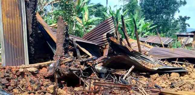 Débris d’une maison entièrement brûlée, avec des tôles tordues et noircies au sol, entourée de végétation.