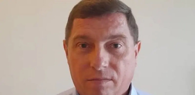 Portrait d’un homme au visage sérieux, portant une chemise claire, posé devant un fond uni beige.