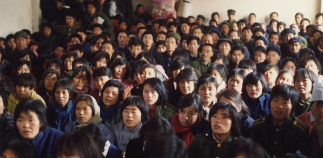 Grande assemblée de personnes assises serrées dans une salle, écoutant attentivement, vêtues de manteaux et uniformes sombres.
