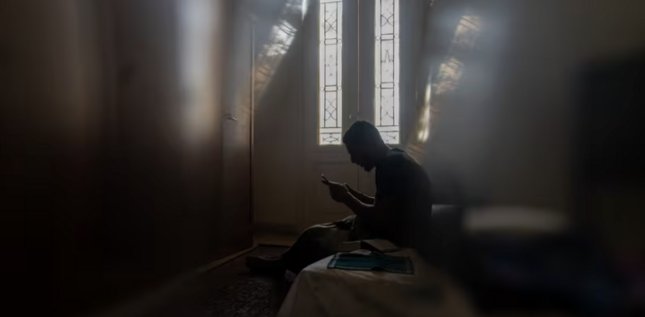Homme assis seul sur un lit dans une pièce sombre, lisant sur son téléphone à la lumière d’une fenêtre étroite.