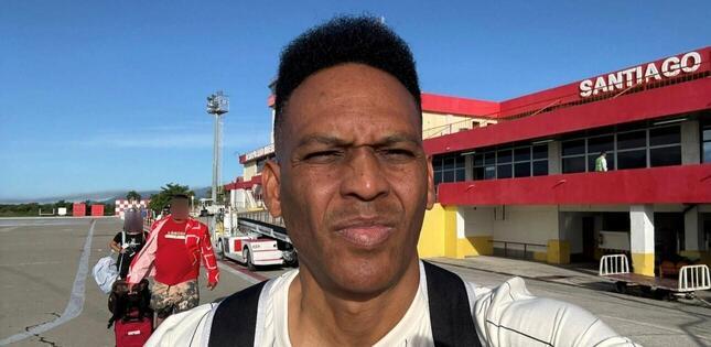 Homme debout sur le tarmac d’un aéroport, le soleil sur le visage, avec le terminal rouge et jaune portant l’inscription « Santiago » en arrière-plan.