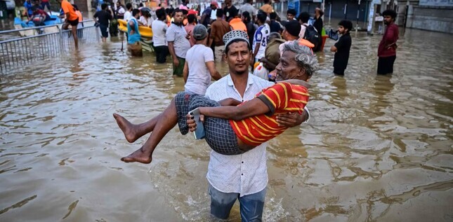 Homme portant un autre homme dans ses bras au milieu d’une rue inondée, entouré de personnes marchant dans l’eau.