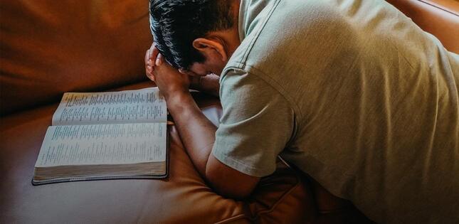 Homme agenouillé, la tête penchée et les mains jointes en prière devant une Bible ouverte sur un canapé.