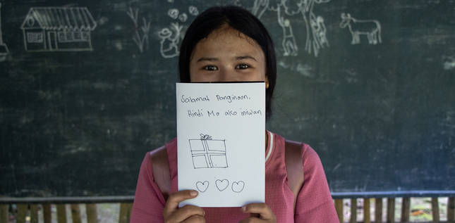Jeune fille souriante tenant devant elle une carte dessinée à la main, avec un message de remerciement et un dessin de cadeau.