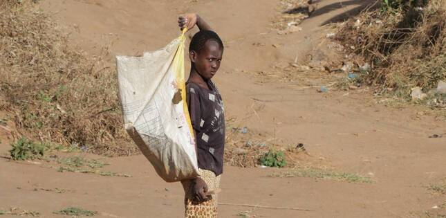 Un jeune garçon pieds nus porte un grand sac sur son épaule et marche sur un chemin en terre dans un village rural.