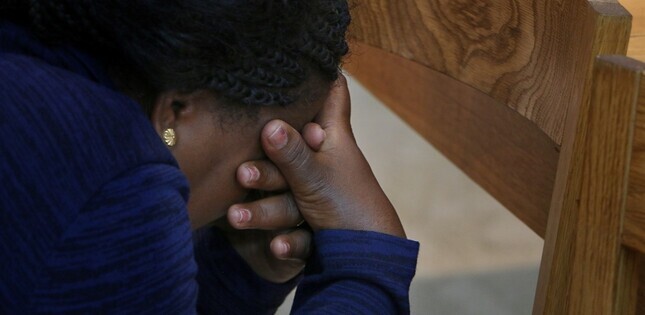 Une femme est assise, la tête baissée et les mains jointes devant son visage, dans une attitude de prière ou de recueillement.