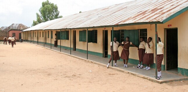 Des écolières en uniforme se tiennent devant un bâtiment scolaire long et simple, dans une cour en terre battue.