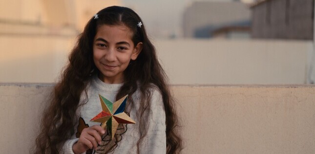 Une jeune fille sourit et tient une étoile colorée en papier devant elle, dans un environnement urbain.