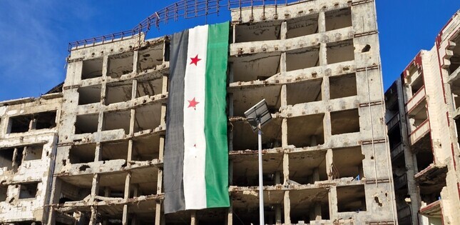 Un bâtiment en ruines porte un grand drapeau syrien, accroché à sa façade dans une rue dévastée.