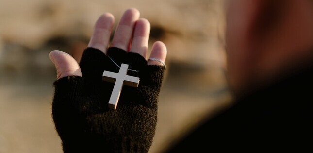 Une main gantée tient une petite croix en métal, suspendue à une chaîne.