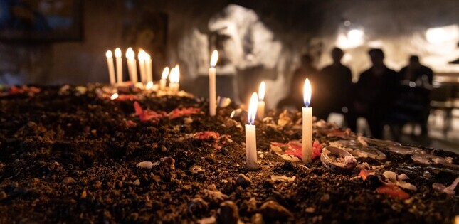 Plusieurs bougies allumées sont posées sur le sol, dans un lieu sombre où des personnes se tiennent en arrière-plan.