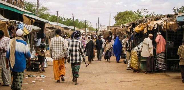 Des personnes marchent dans une rue animée bordée d’étals de marché, dans un environnement urbain modeste.