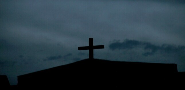 Silhouette d’une croix au sommet d’une église, se détachant sur un ciel sombre et nuageux.