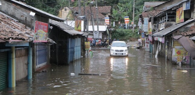 Une rue commerçante est inondée, avec de l’eau recouvrant la chaussée et une voiture avançant lentement entre les bâtiments.