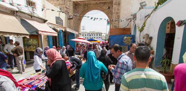 Des gens se déplacent dans une rue animée, avec des étals de marché et des parasols colorés, sous une arche ancienne.