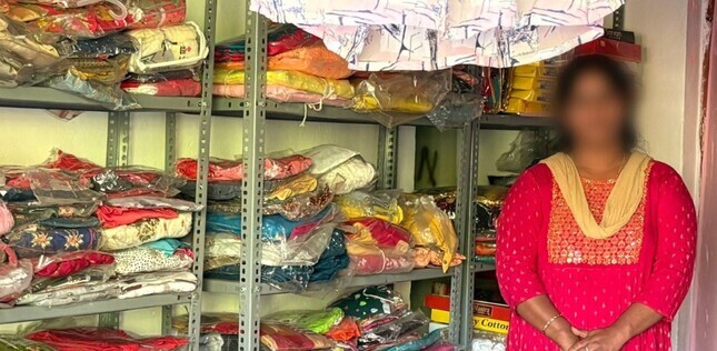 Une femme se tient dans une petite boutique de vêtements, devant des étagères remplies de tissus et d’habits pliés.