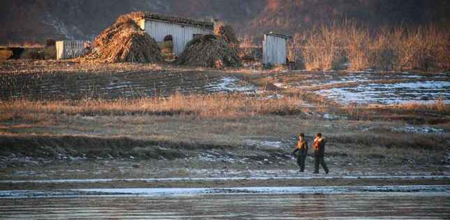 Deux hommes marchent le long d’un cours d’eau dans un paysage rural froid, avec des bâtiments simples et des tas de végétation en arrière-plan.
