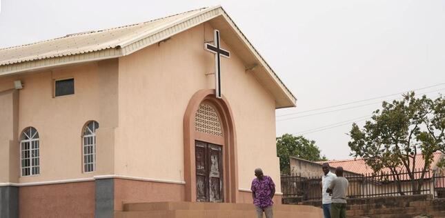 Une petite église avec une croix au-dessus de l’entrée se dresse dans un quartier résidentiel, avec quelques hommes à proximité.