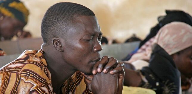 Un jeune homme est assis les yeux fermés, les mains jointes devant son visage, dans une attitude de prière lors d’un rassemblement.