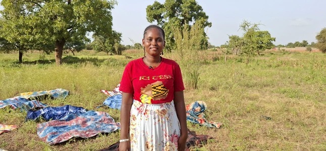 Une jeune femme se tient dans un champ verdoyant, avec des tissus étendus au sol derrière elle.