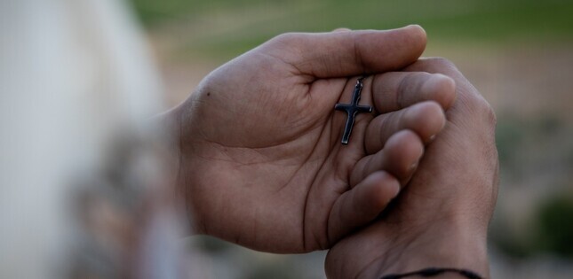 Gros plan sur deux mains jointes tenant une petite croix noire, dans un geste de prière. L’arrière-plan est flou, avec des tons verts et naturels.