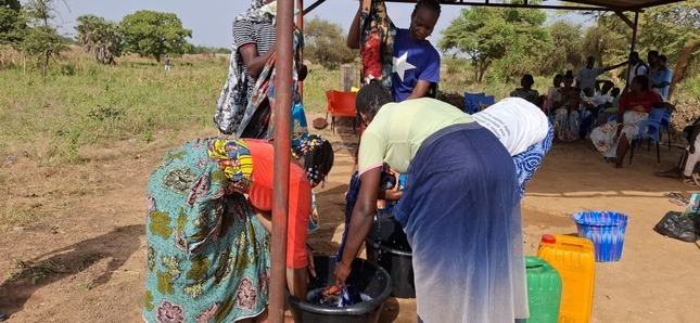 Des femmes teignent des tissus dans des bassines d’eau, sous un abri en plein air, dans un environnement rural.