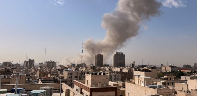 Vue d’une ville avec d’épais panaches de fumée s’élevant derrière des immeubles, sous un ciel dégagé.