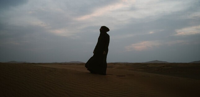 Silhouette d’une femme vêtue de noir marchant sur des dunes de sable, sous un ciel nuageux au crépuscule.