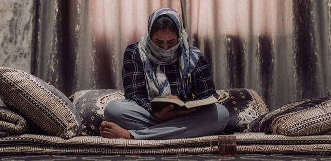 Femme assise sur un canapé, la tête baissée, lisant un livre dans une pièce sombre.