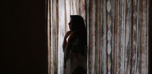 Vue d’une femme vêtue d’un voile, debout près de fenêtres aux rideaux épais, plongée dans une réflexion silencieuse.