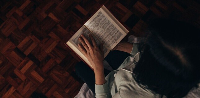 Femme lisant un livre, ses mains effleurant les pages sur un parquet en bois.