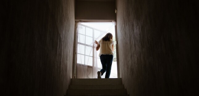 Silhouette d’une femme montant un escalier sombre vers une porte ouverte et lumineuse.