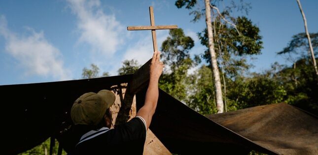 Un homme installe une croix en bois sur le toit d’un bâtiment, au milieu de la végétation.
