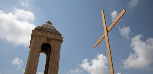 Une grande croix en bois dressée à côté d’une tour d’église sous un ciel nuageux.