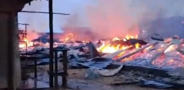 Des habitations en flammes après une attaque, avec d’épaisses fumées s’élevant au-dessus des décombres.