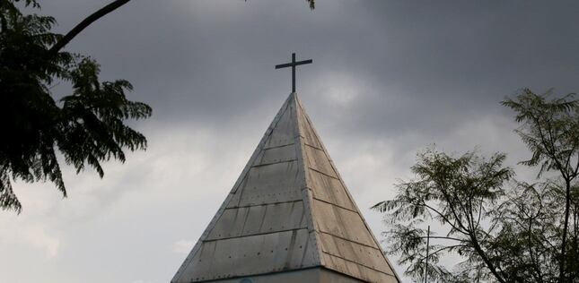 Clocher d’une église surmonté d’une croix, entouré d’arbres sous un ciel nuageux.