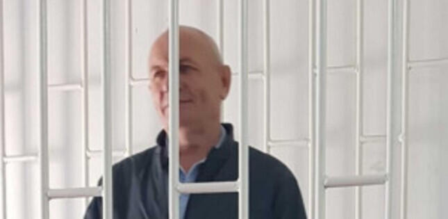 Un homme se tient derrière des barreaux de prison, dans une pièce aux murs clairs.