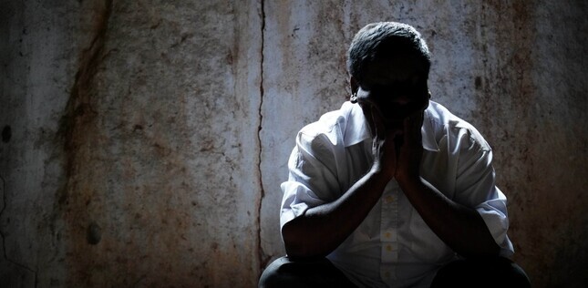 Un homme assis dans l’ombre, le visage enfoui dans ses mains, dans une pièce aux murs bruts.