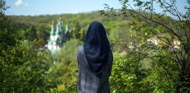 Une femme de dos, voilée, contemple un paysage verdoyant où se dresse une église au loin.