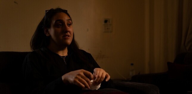 Portrait d’une jeune femme assise dans une pièce sombre, éclairée par une lumière tamisée.