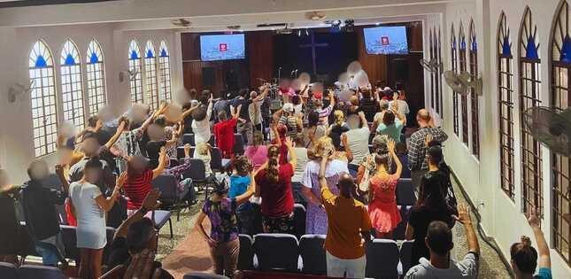 Vue d'une assemblée dans une salle avec une croix au fond, plusieurs personnes levant les mains.